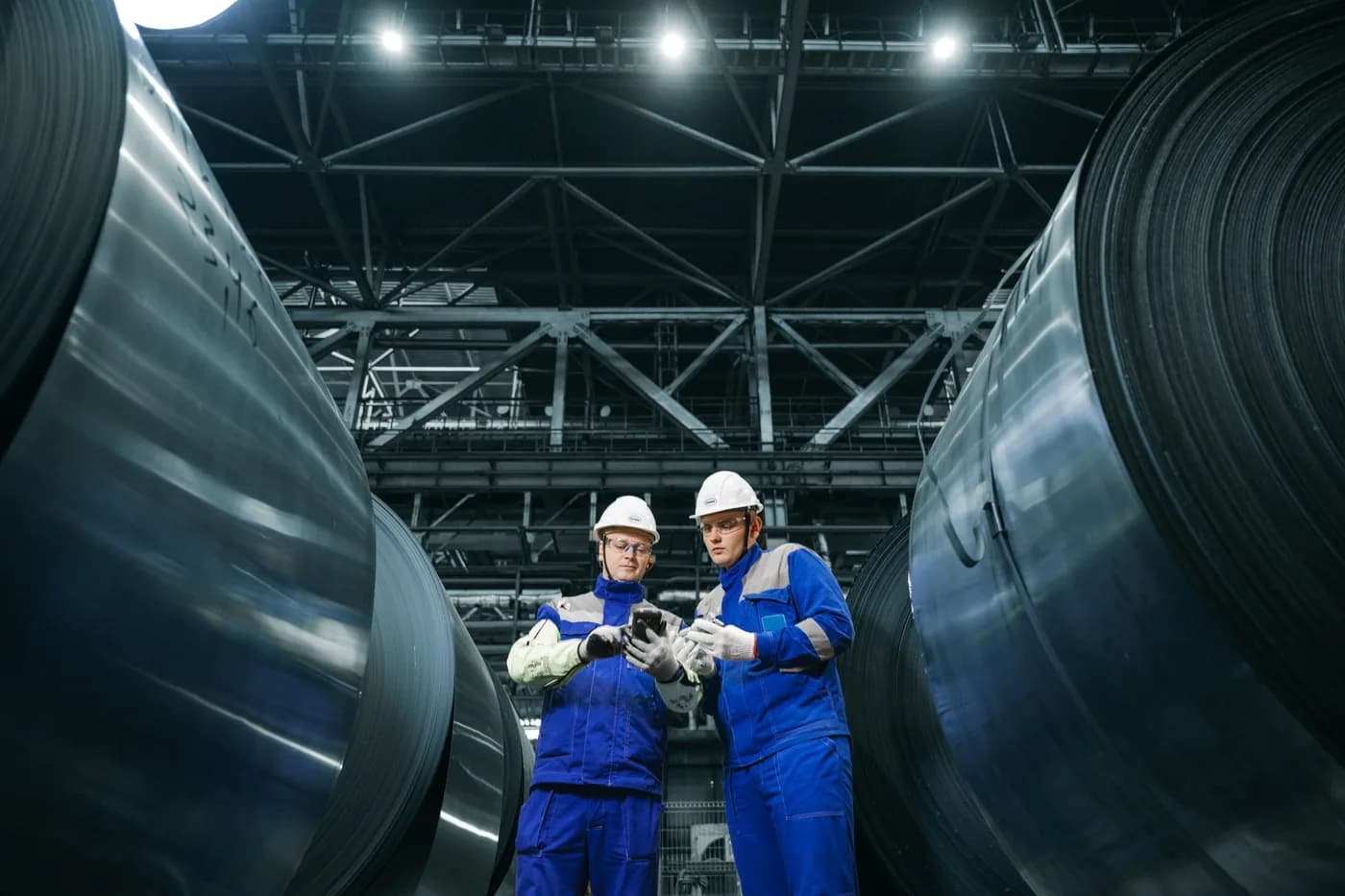 Two workers in hard hats inspecting rolled steel in an industrial hall